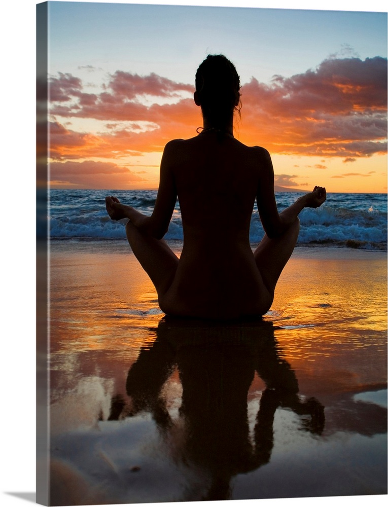 Woman meditating and doing yoga on the wet sand as the surf breaks in front of her and the sun setting creating a silhouet...