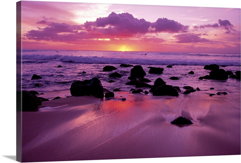 Several dark rocks dot the beach among shallow waves at twilight as purple clouds loom overhead.