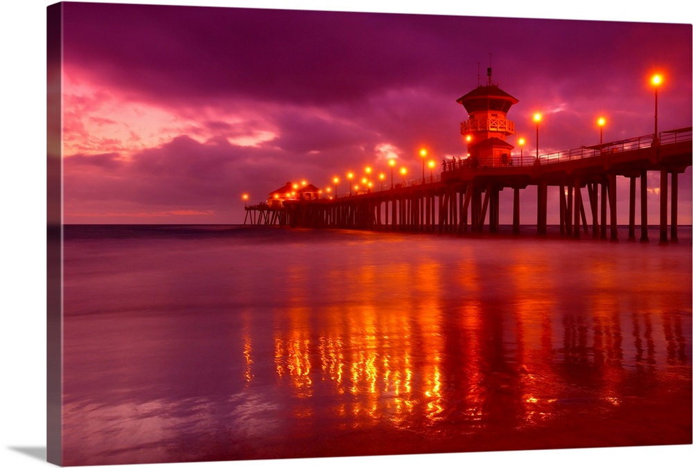 Big horizontal photograph of a lit pier reflecting in the water near Huntington beach at dusk, beneath a vibrant, cloudy sky.