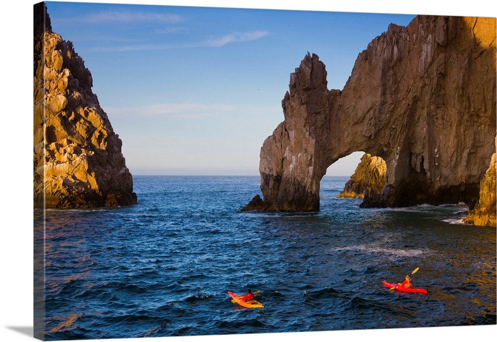 Two people in kayaks paddle in the ocean near tall rock arch formations.