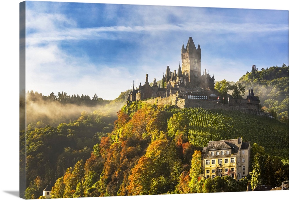 Large medieval castle on a colorful tree hillside with fog, blue sky, and cloud, Cochem, Germany.