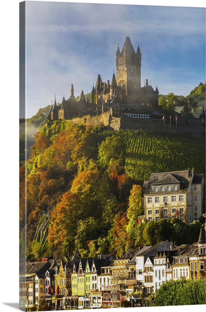 Large medieval castle on top of a colorful treed hillside with fog, blue sky, cloud, and village below, Cochem, Germany.