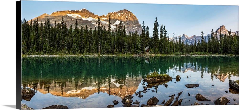 Panorama of mountain range reflecting in an alpine lake with the glow of warm early morning light and blue sky; British Co...