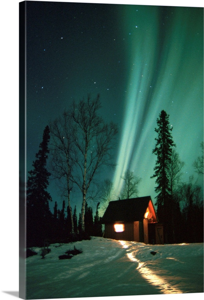 A photograph of the Aurora Borealis wisps in starry sky above a small cabin in the snow.