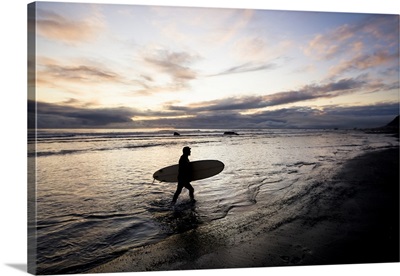 Silhouette Of A Surfer Carrying A Surfboard Back To Shore At Sunset; Alaska