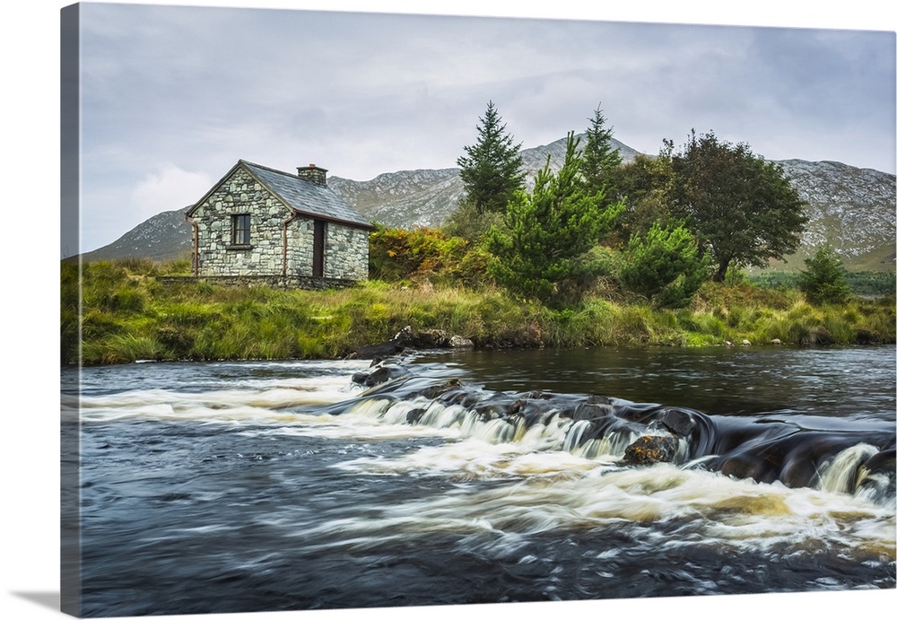 Small stone fisherman's hut on the banks of a small river with mountains in the background on a cloudy day; Connemara, Cou...