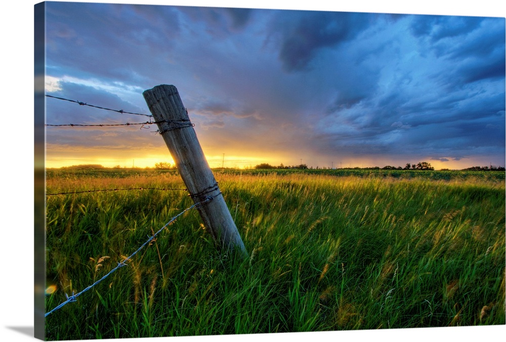 Photograph of wooden fence with barbwire in a field under dark storm clouds.
