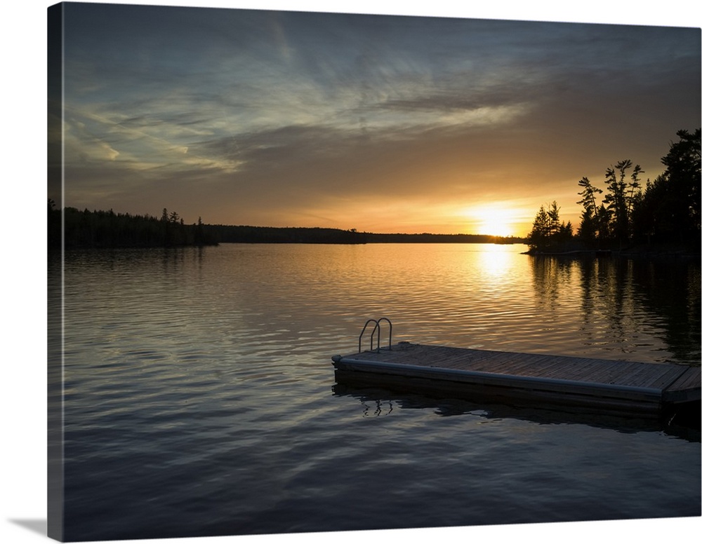 The sun setting over a tranquil lake and silhouetted trees along the shoreline with a dock and ladder in the foreground; L...
