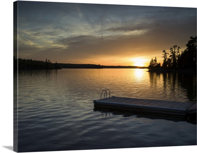 The sun setting over a tranquil lake and silhouetted trees with a dock ...