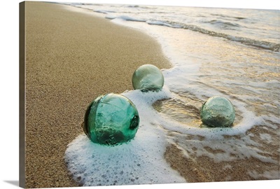 Three Glass Fishing Floats Roll On The Sandy Shoreline With Ripples Of Water And Seafoam