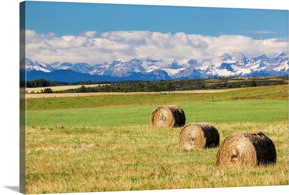 Three hay bales in a field with mountains in the background slightly snow covered and cloudy with blue sky, Alberta, Canada.