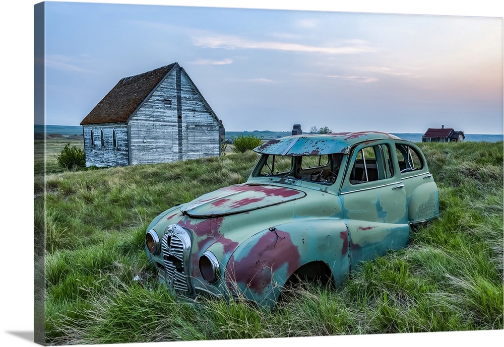 Vintage car sitting in the overgrown grass in a field with old buildings on a farmstead; Saskatchewan, Canada