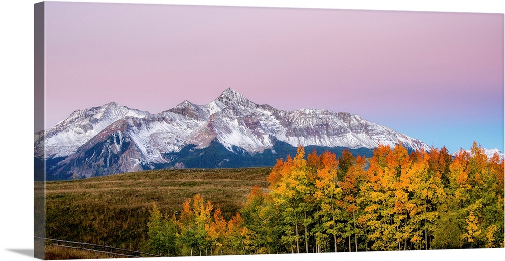 Landscape photograph of a snowy mountain range with colorful Fall trees in the foreground and a cotton candy sky above.