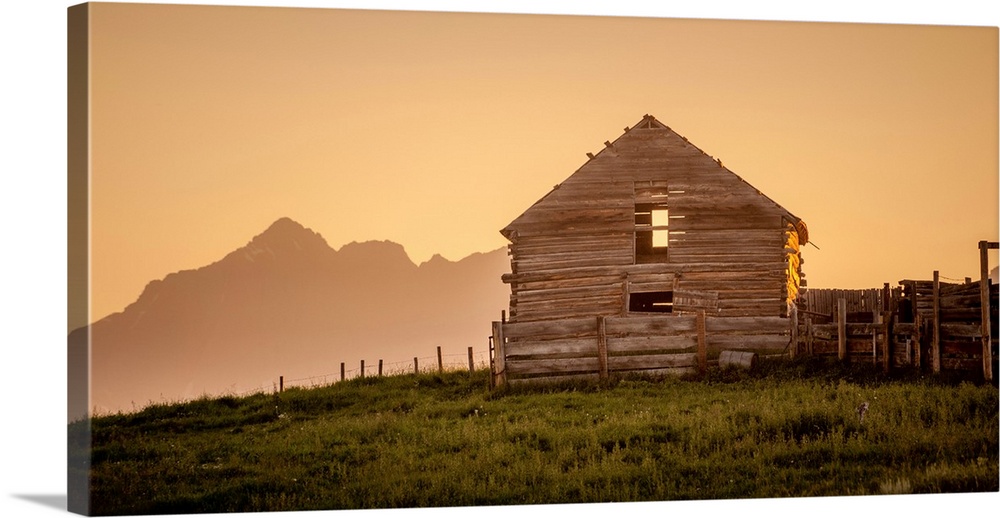 Warm photograph of an old wooden barn on a hilltop with a silhouette of mountains in the background.