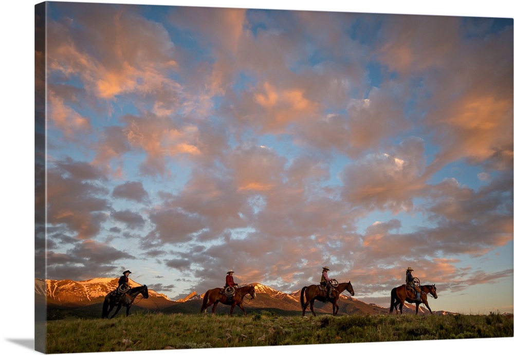 Sunset photograph with four people on horseback riding through a valley in a line with snow capped mountains in the backgr...