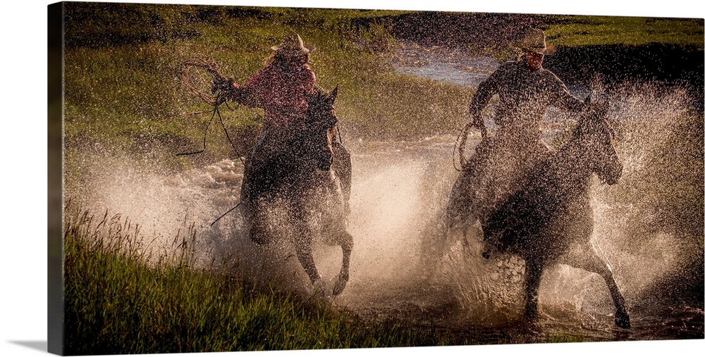 Photograph of two cowgirls splashing through a steam on horseback with their lassos out.