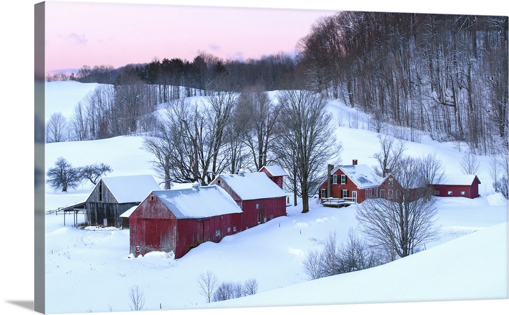 Photograph of a snowy rolling hills with red buildings and bare Winter trees at sunrise.