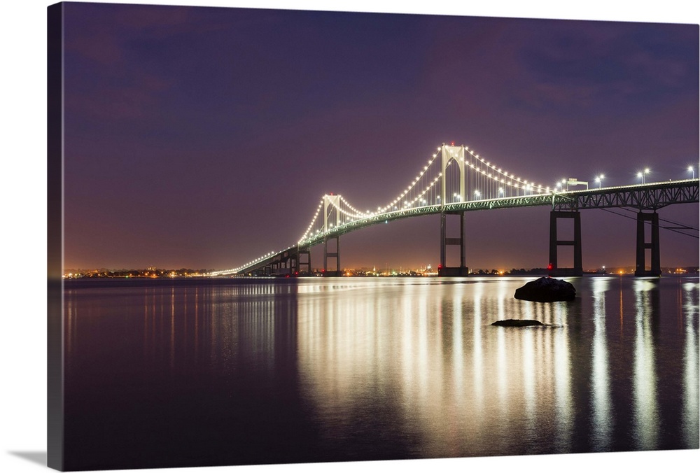 A photograph of a large suspension bridge seen lit up at night casting long reflections on the water below.