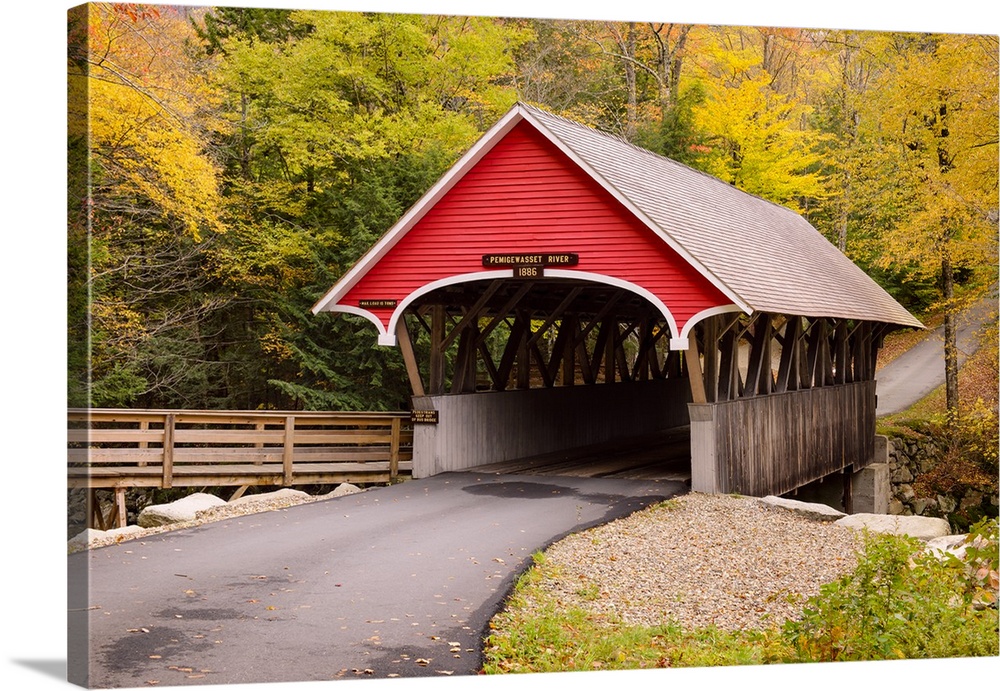 Photograph of a bright red covered bridge with a road leading up a hill lined with yellow, orange, and green Fall trees.