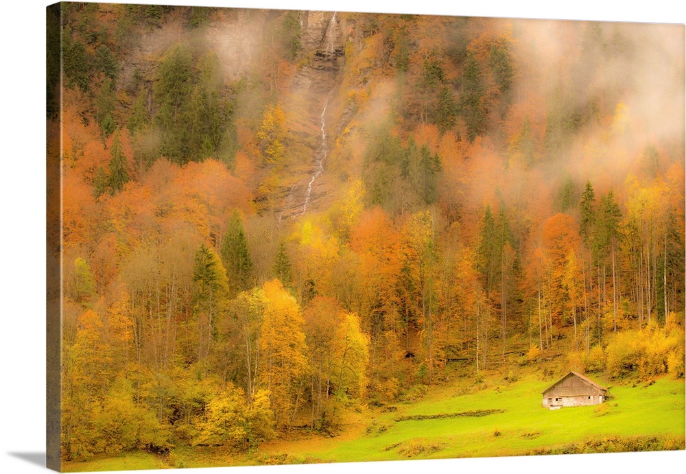 Landscape photograph of colorful Autumn trees on the side of a foggy mountain with a small cabin and waterfall.