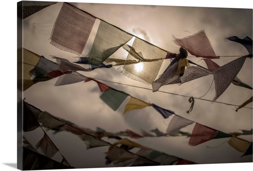 Photograph of colorful, worn prayer flags blowing in the wing with the sun shining through the clouds in the background.