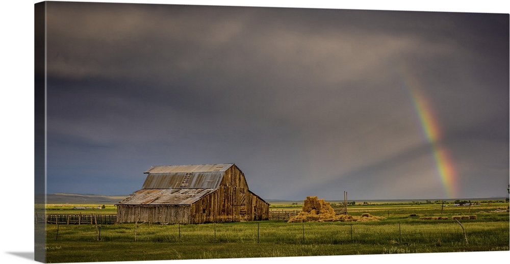 A photograph of an old barn in a wilderness landscape with a rainbow peaking out from clouds in the distance.