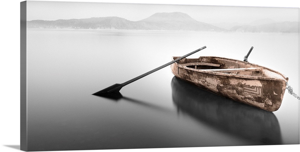 An artistic photograph of a rowboat with no passenger sitting on flat water with a mountainous landscape seen in the foggy...