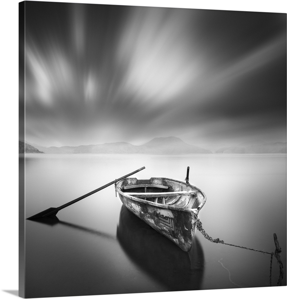 An artistic black and white photograph of a lone and unattended rowboat on a still watery surface under a motion blurred sky.