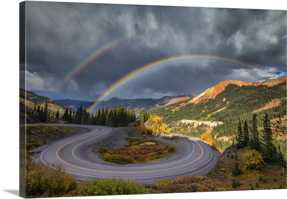 A double rainbow in the sky over a bend in the road in the mountain wilderness.