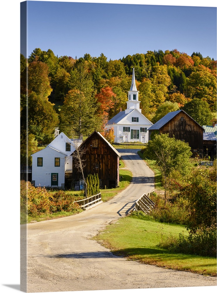 Photograph of a gravel road leading up the hill to a white church surrounded by various wooden buildings and colorful Fall...