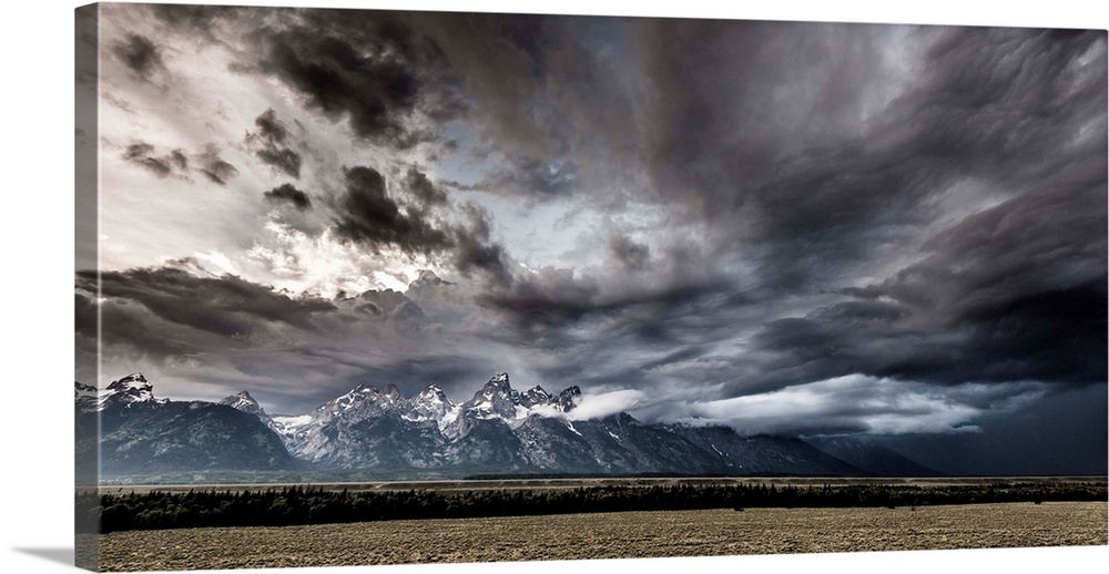 Landscape photograph of a field in front of snow capped mountains with a dramatic stormy sky above.