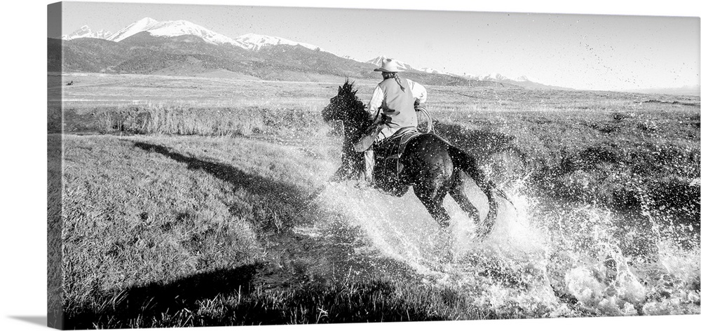 Action photograph of a cowgirl splashing across a river on horseback with snow capped mountains in the distance.