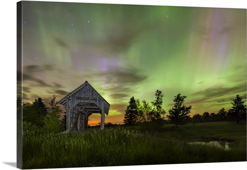 Long exposure photograph of the Northern Lights at night in an open field with a covered bridge.