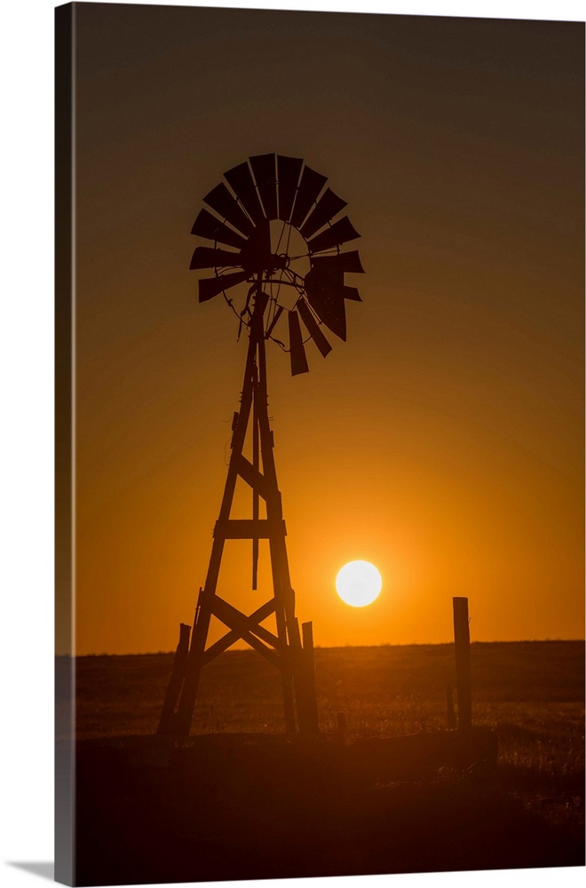 Silhouette photograph of a windmill at sunset with the round sun sitting right above the horizon line.