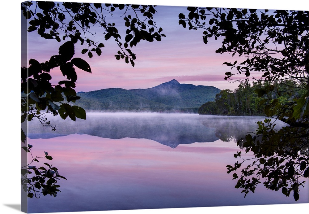 Landscape photograph of a mountain view reflecting onto a foggy lake with a pink and purple sunset framed by leafy tree br...