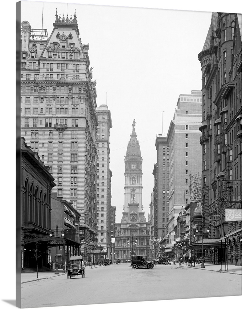 This is a vintage photograph in black and white of downtown Philadelphia. Tall buildings line the street that has cars dri...