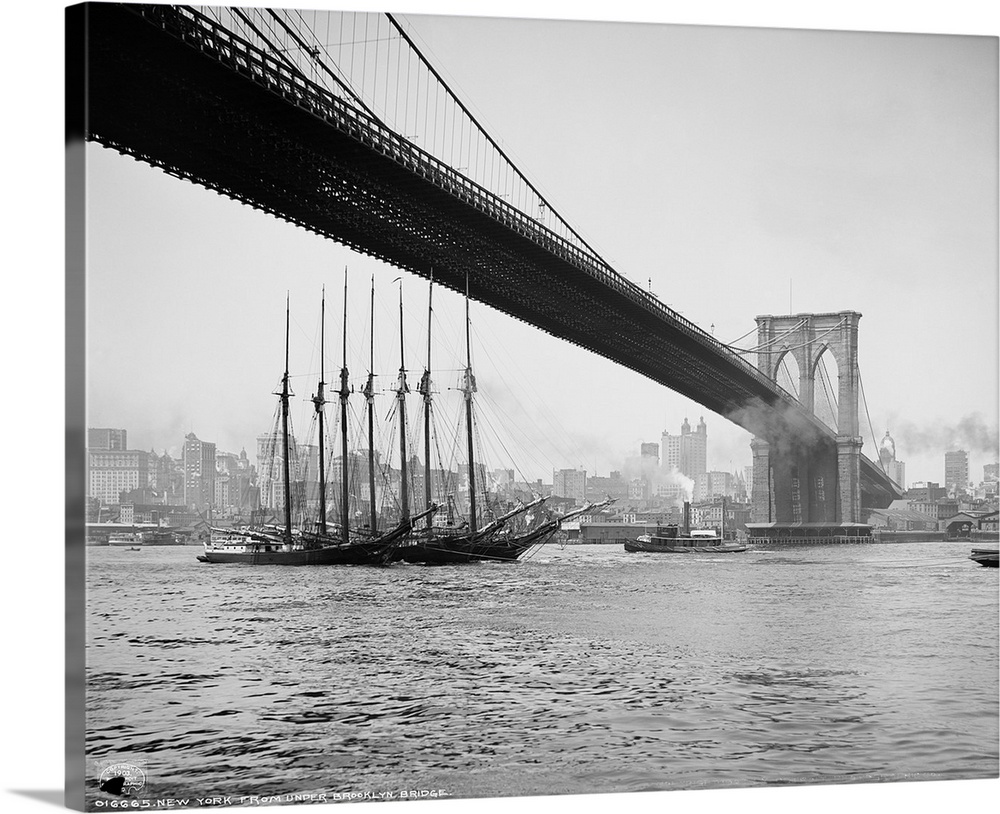 Horizontal, vintage photograph of an old ship passing beneath the Brooklyn Bridge, along the East River, the New York City...