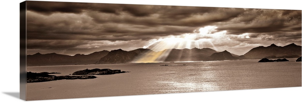 Panoramic photo, in a sepia tone, of the sun streaming through a heavy clouded sky over a body of water with mountains in ...