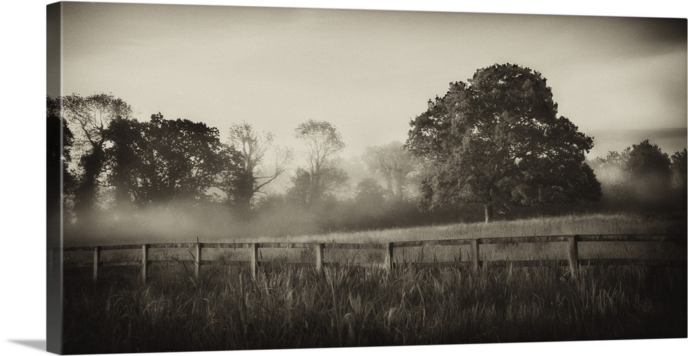 A horizontal photograph of a country field with a wooden fence and mist over trees in the distance.