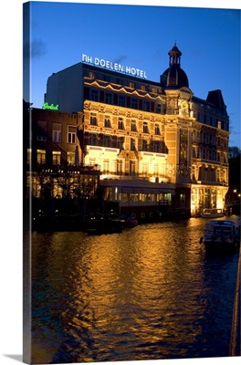 Doelen Hotel with lights on at night along the Amstel River in Amsterdam, Netherlands