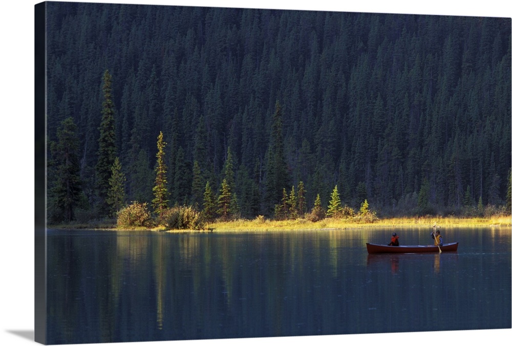 Two people paddle in a canoe in the early morning light, with lush forest behind them, on Waterfowl Lake, in Banff Nationa...
