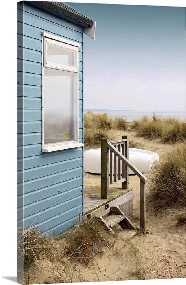 View of the side of a blue wooden beach hut with a wooden terrace, looking towards the coast/beach. A white upturned boat ...