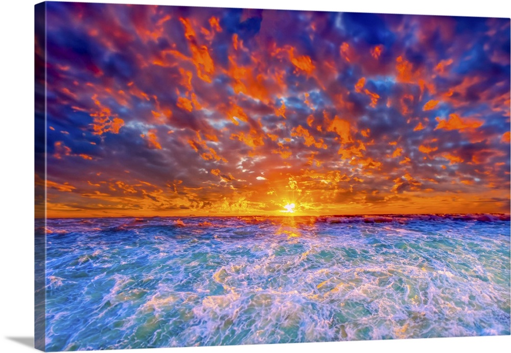 A fiery red, orange, purple, and blue sunset explodes a into colorful sunset. Gold crested waves dance in the ocean below.