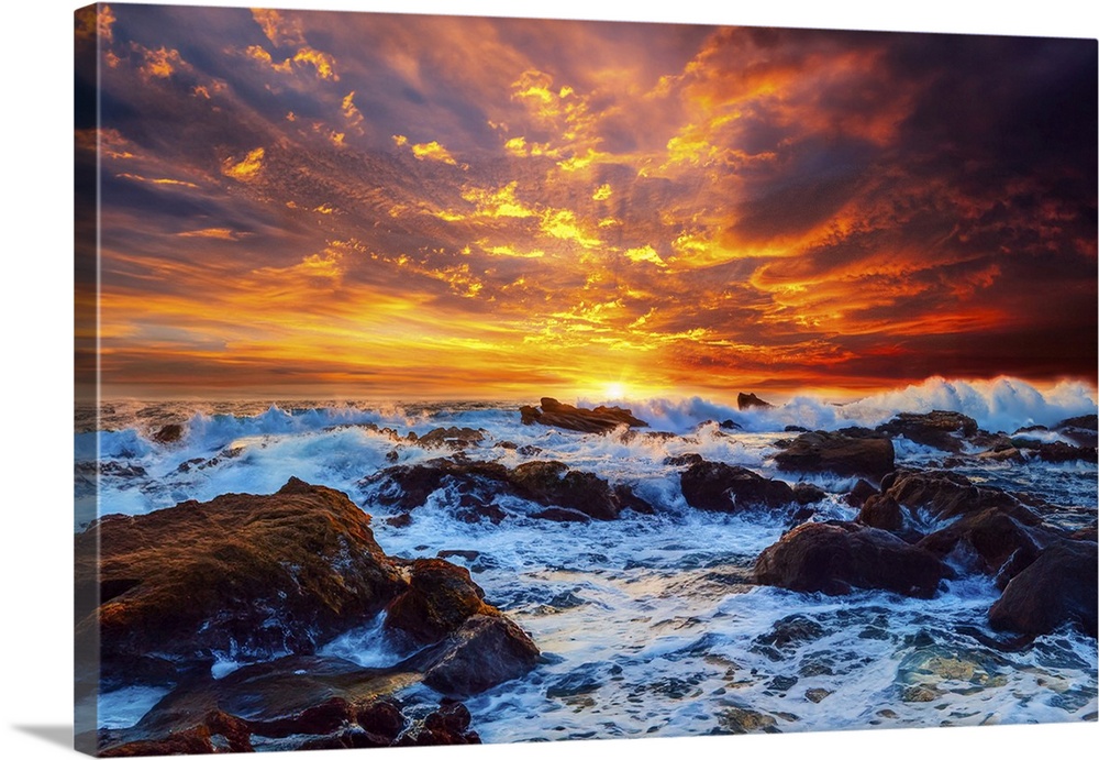 A bright fiery red sky illuminates crashing waves on the rocks in the ocean.