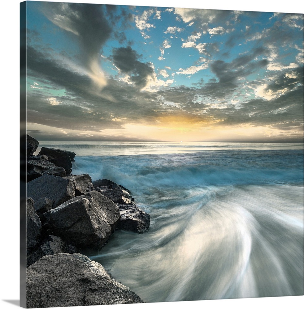 Long exposure photograph of ocean waves crashing on a rocky beach shore with a dramatic sky.