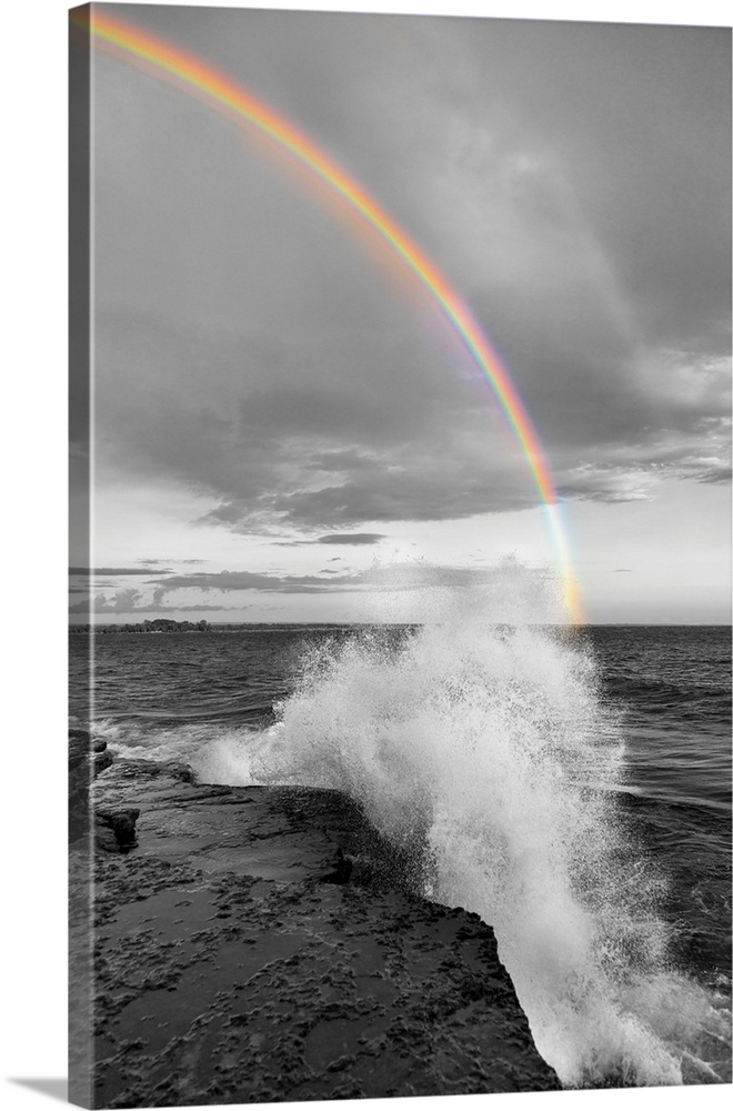 Black and white photograph of a waves crashing on a rocky shore with a colorful rainbow over the water.