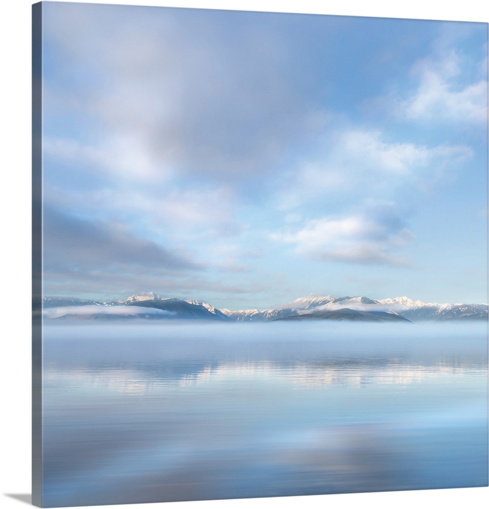 Square photograph of a snow capped mountain range in the distance with fluffy clouds above reflecting onto the ocean.