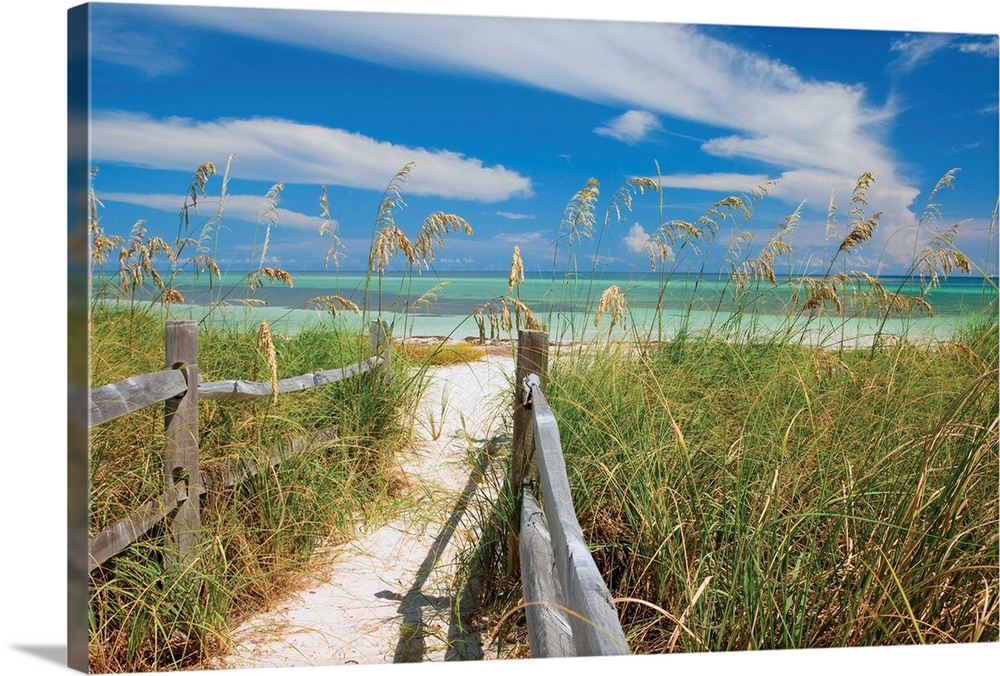 Photograph of a sandy path lined with a wooden fence and beach grass leading to a sandy beach with clear, teal waters and ...