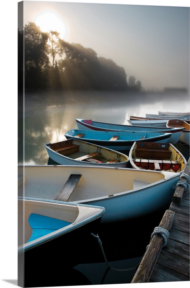Photograph of a cluster of boats docked on a lake with beautiful morning light peering through the foggy trees.