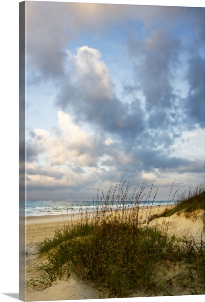 Vertical photograph of grassy dunes in front of the beach, beneath a sky of billowing clouds as the sun rises.